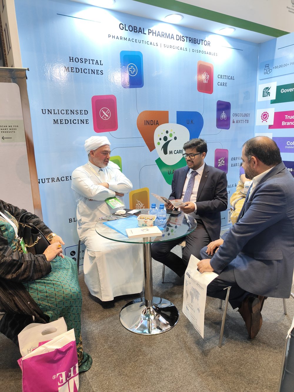 M Care Arab Health 2025 booth, "Global Pharma Distributor" wall graphic, meeting in progress at round table.